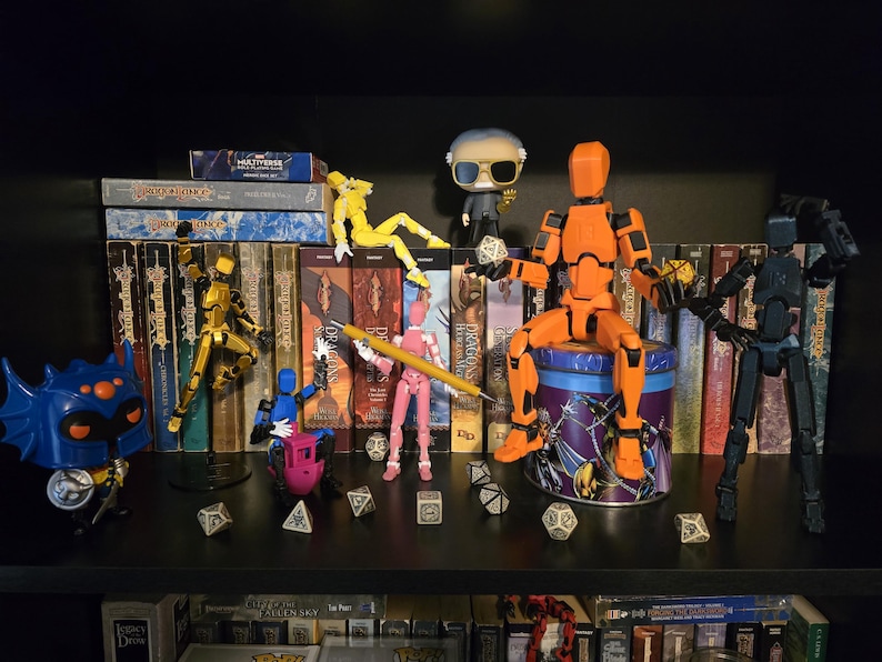 A group of Dummy 13 articulated figures enjoying a session of Dungeons and Dragons with a back drop of Dragonlance novels and Funko Pop dolls of Stan Lee and Warduke. The pink figure holds a pencil, while the orange Titan figure gets ready to roll a d20. A multicolored figure rides on a 3d printed "Benchy."