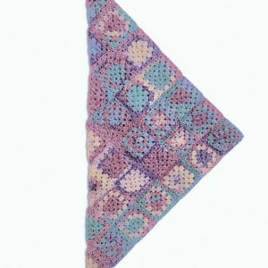 May include: A triangular, handmade crochet shawl in pastel colors. The shawl features a repeating pattern of squares in shades of lavender, light blue, and cream. The soft, textured fabric is displayed against a white background.