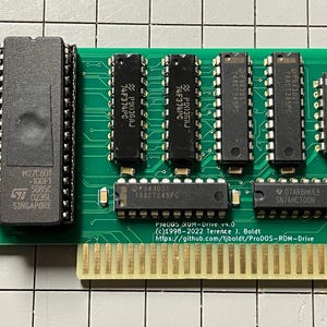 Puede incluir: Una placa de circuito verde con múltiples chips de circuito integrado negros y un conector dorado. La placa tiene texto, incluyendo "PRODOS ROM-Drive v4.0" y otras marcas técnicas. Los chips están dispuestos en filas.