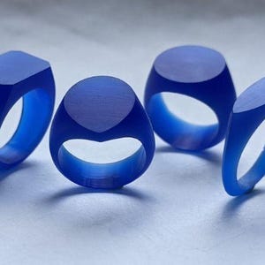 Puede incluir: Cuatro anillos azules con diferentes formas geométricas. Uno tiene la parte superior rectangular, otro ovalada, un tercero circular y el último ovalada inclinada. Los anillos están hechos de un material translúcido.