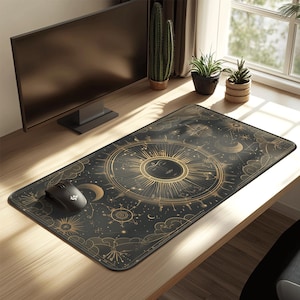 Op de afbeelding: Zwart rechthoekig bureau muismat met een gouden hemels ontwerp. Het ontwerp toont een zon, maan, sterren en astrologische symbolen. Een zwarte computermuis ligt aan de linkerkant van de mat.