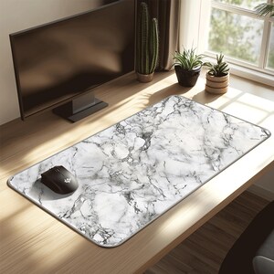May include: A rectangular mouse mat with a white and grey marble design, with a black mouse on the left. The mat sits on a wooden desk, with a computer monitor and plants visible in the background.