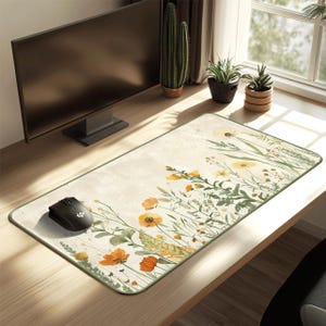 Op de afbeelding: Rechthoekige muismat met een bloemenontwerp. De mat heeft een crèmekleurige achtergrond met oranje en gele bloemen, groene bladeren en een groene rand. Een zwarte computermuis ligt aan de linkerkant.