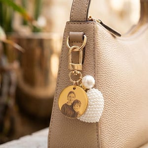 Könnte beinhalten: Ein goldfarbener Schlüsselanhänger mit einem runden Anhänger, der ein Foto von zwei Personen zeigt. Der Schlüsselanhänger ist an einer beigen Handtasche befestigt. Eine weiße Perle und ein herzförmiger Cluster aus kleinen weißen Perlen sind ebenfalls am Schlüsselanhänger befestigt.