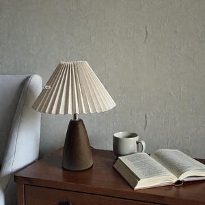 May include: A beige pleated lampshade sits atop a dark brown wooden lamp base. The lamp is on a wooden nightstand next to a gray mug and an open book. The background features a textured gray wall and a white armchair.