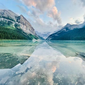 May include: A scenic landscape featuring a calm lake reflecting the sky and surrounding mountains. The water's surface mirrors the clouds and the peaks, creating a symmetrical image. The mountains are covered in green trees and rocky cliffs.