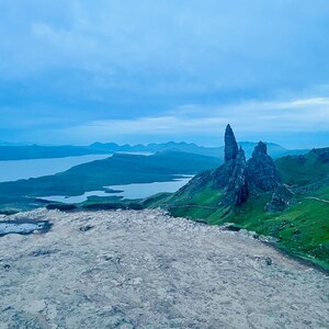 Puede incluir: Un paisaje escénico con un primer plano rocoso, verdes colinas y una masa de agua bajo un cielo azul nublado. Formaciones rocosas irregulares se alzan desde las verdes colinas. Un sendero sinuoso atraviesa el paisaje.
