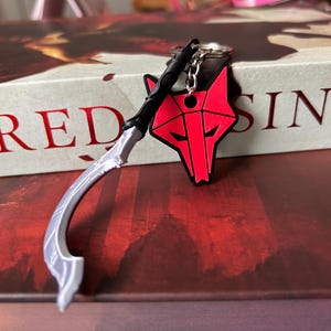 May include: A keychain featuring a silver-colored curved blade with a black handle and a red wolf head emblem. The wolf head has black outlines and a geometric design. The keychain is attached to a metal ring by a short silver-colored chain.
