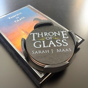 Peut inclure: Ensemble de dessous de verre noirs avec l'inscription "THRONE OF GLASS" et "SARAH J. MAAS" en blanc, maintenus par un support noir. Les dessous de verre reposent sur un livre du même titre.