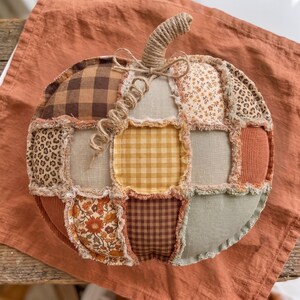 Può includere: Una zucca rotonda in tessuto con un design patchwork composto da vari tessuti, tra cui quadri marroni, stampa leopardata, quadretti gialli, motivi floreali e verde salvia tinta unita. La zucca ha un picciolo in corda di iuta.