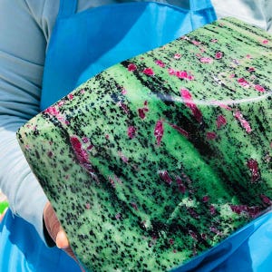 May include: A large, polished block of zoisite mineral featuring a vibrant green base color, interspersed with dense black mineral inclusions and scattered, irregular bright pink ruby crystal formations throughout the stone surface.