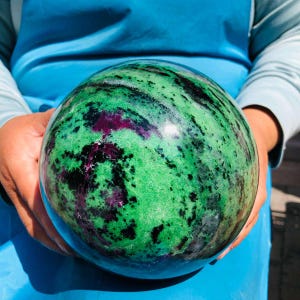 May include: A polished spherical stone featuring a vibrant green base color with prominent black mineral inclusions and deep magenta-colored patches throughout the surface, showcasing a smooth, reflective texture and a rounded, orb-like shape.