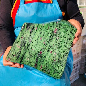 May include: A large, polished slab of zoisite mineral featuring a vibrant green base color with prominent black hornblende inclusions and scattered, small, opaque magenta-colored ruby crystals throughout the irregular, angular stone surface.