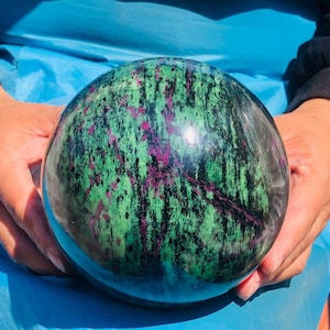May include: A polished spherical stone featuring a vibrant mix of deep green, black, and magenta-pink mineral inclusions. The surface is smooth and reflective, showcasing a natural marbled pattern with distinct dark streaks and bright pink patches.