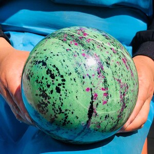 May include: A polished spherical stone featuring a vibrant green base color with scattered magenta-pink inclusions and dark black mineral streaks throughout the surface. The sphere has a smooth, reflective texture and a natural, variegated pattern.