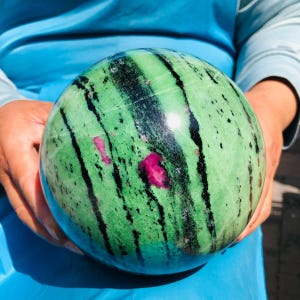 May include: A polished spherical stone featuring a vibrant green base color with prominent black streaks running vertically across the surface. The sphere also displays small, irregular patches of deep magenta or pink mineral inclusions.