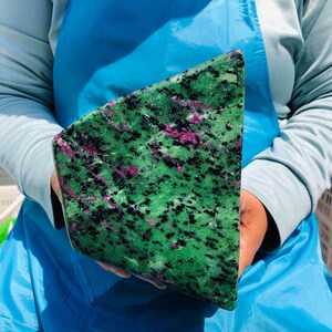 May include: A large, triangular-shaped slab of polished zoisite mineral featuring a vibrant green base color, speckled with dark black hornblende inclusions and distinct, irregular patches of bright magenta-colored ruby crystals throughout.