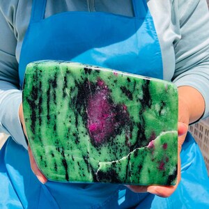 May include: A polished slab of zoisite mineral featuring a vibrant green base color with prominent black hornblende inclusions and a large, central patch of deep magenta ruby crystal. The stone has a smooth, flat surface with natural textures.