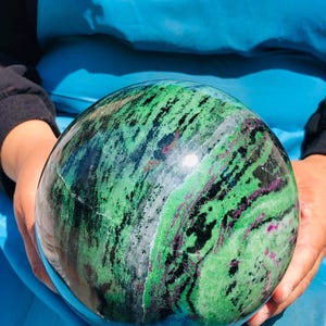 May include: A polished spherical stone featuring a vibrant mix of bright green, deep black, and magenta-pink mineral inclusions. The surface displays a smooth, glossy texture with swirling patterns of color throughout the entire round object.