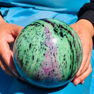 May include: A polished spherical stone featuring a vibrant mix of light green, deep magenta, and black mineral inclusions. The surface is smooth and reflective, displaying natural patterns of contrasting colors throughout the round mineral specimen.