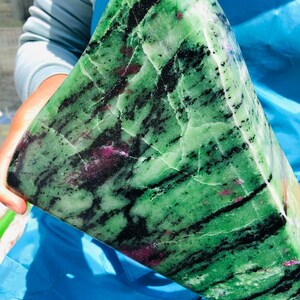 May include: A large, polished zoisite mineral specimen featuring a vibrant green base color with prominent black hornblende inclusions and scattered, opaque magenta-colored ruby deposits throughout the crystalline structure of the stone.