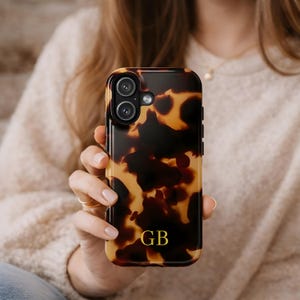 Puede incluir: Funda de teléfono con diseño de carey y las iniciales doradas "GB". La funda tiene un acabado brillante y una abertura para una cámara de doble lente. La funda del teléfono se sostiene en una mano.