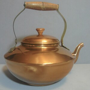 May include: A copper kettle with a wooden handle and a brass spout. The kettle has a shiny, polished finish.