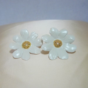 May include: A pair of white flower-shaped earrings with yellow centers. The earrings are made of a smooth, glossy material.