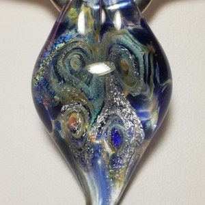 May include: A dichroic glass pendant with a silver bail. The pendant is a teardrop shape with swirling patterns in shades of blue, silver, yellow, and orange. The glass has a reflective, iridescent quality.