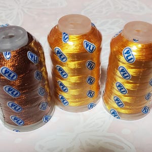 May include: Three spools of metallic thread in shades of bronze and gold. Each spool is wrapped in clear plastic with a blue and white label that reads "YLI" and "174". The thread is tightly wound on clear plastic spools.