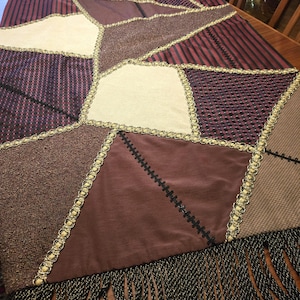 May include: A rectangular throw blanket featuring a patchwork design of various fabrics in shades of burgundy, brown, and cream. The segments are joined by decorative gold-toned braided trim and black stitching, finished with long black fringe.