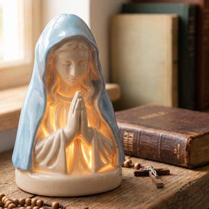Puede incluir: Una figura de cerámica de una persona con las manos juntas en oración, que lleva un velo azul claro. La figura está iluminada desde el interior, proyectando un brillo cálido a través del material translúcido, con un acabado liso y brillante.