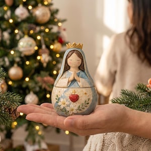 Pode incluir: Uma pequena estatueta arredondada de uma figura com cabelo castanho, um véu azul e uma coroa dourada. As mãos estão postas em oração. A base apresenta um coração vermelho com uma cruz e flores brancas sobre um fundo creme.