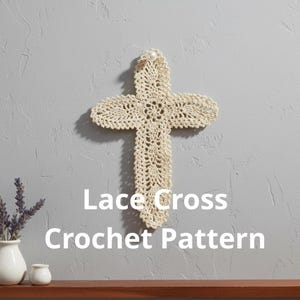 Può includere: Una croce in pizzo all'uncinetto color crema con un accento di perla in alto, appesa a una parete grigia. La croce è un oggetto decorativo, forse per scopi religiosi o di arredamento. Il testo sull'immagine recita "Lace Cross Crochet Pattern."