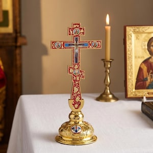 Peut inclure: Une croix orthodoxe sur pied avec une finition en émail rouge, des motifs en filigrane doré, de petites icônes religieuses circulaires et un crucifix central. La croix est fixée sur une base en métal doré poli avec des gravures.