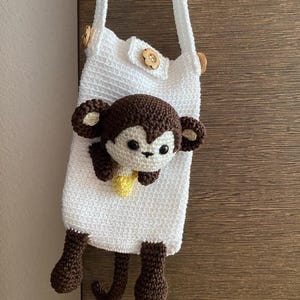 Può includere: Una borsa all'uncinetto bianca fatta a mano con un'applicazione a forma di scimmia marrone. La scimmia ha viso, orecchie e arti marroni, con una banana gialla. La borsa ha una tracolla e chiusure con bottoni in legno. La borsa è alta circa 20 cm.