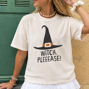 Op de afbeelding: Een ivoorkleurig T-shirt met korte mouwen en een afbeelding van een zwarte heksenhoed met een oranje en zwarte stippenband. Onder de hoed staat de tekst WITCH, PLEEEASE! in zwarte hoofdletters. Het shirt heeft een ronde hals en een losse pasvorm.