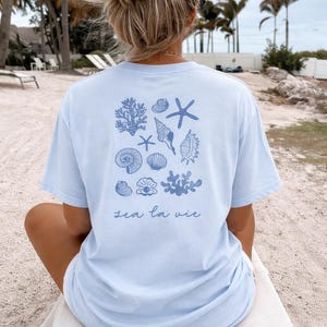 Może przedstawiać: Jasnoniebieska koszulka z nadrukiem muszli i koralowców na plecach. Koszulka ma napis "sea la vie". Projekt zawiera różne muszle i koralowce w niebieskim odcieniu.