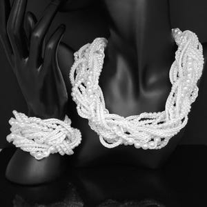 May include: A white pearl necklace and bracelet set displayed on a black mannequin. The necklace is a multi-strand design, and the bracelet is a matching style. The image has a high-contrast, studio lighting effect.