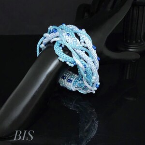 May include: A woven bracelet in shades of blue and white, decorated with small beads. The bracelet is displayed on a black surface. The letters "BIS" are visible in the lower left corner.