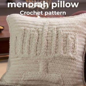 Puede incluir: Cojín de ganchillo color crema con diseño de menorá. El cojín presenta una superficie texturizada y las palabras "Crochet menorah pillow Crochet pattern" en blanco. El cojín está colocado sobre una superficie granate.