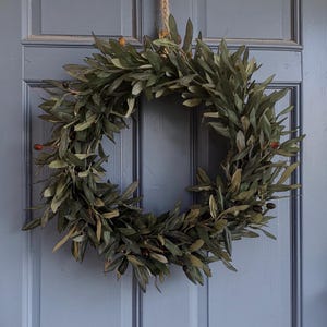 May include: A circular olive branch wreath with green leaves and small, dark berries. The wreath is suspended by a natural fibre rope against a blue door. A decorative accent for the home.