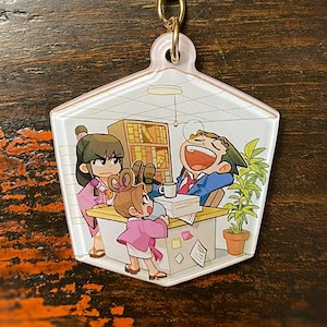 Może przedstawiać: Złoty brelok do kluczy z kreskówkową ilustracją trzech osób w biurze. Brelok ma złoty łańcuszek i przezroczysty, sześciokątny urok. Urok przedstawia mężczyznę w niebieskim garniturze śmiejącego się.