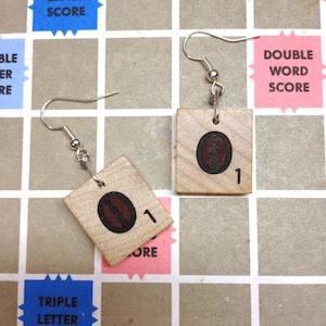 May include: Handmade wooden earrings featuring Scrabble tiles. Each earring has a light wood square tile with a brown oval design and the number 1. The earrings hang from silver-colored fish hook ear wires.