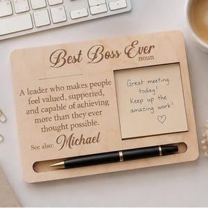 May include: Wooden desk organiser with the text "Best Boss Ever" and a definition. Includes a notepad with a handwritten message, a black pen, and a slot for the pen. The organiser is on a white surface.