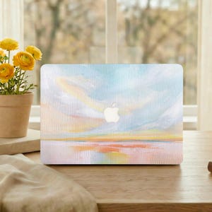 May include: A laptop cover featuring an abstract landscape painting in pastel colors. The design includes a sky with blue and yellow hues, a horizon line, and a reflective surface. The Apple logo is visible.