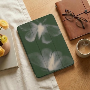 Pode incluir: Uma capa de tablet verde escuro com um design floral branco. A capa está aberta, revelando a tela. A imagem também inclui um diário de couro marrom, óculos e uma xícara de café, sugerindo um espaço de trabalho ou uma mesa.