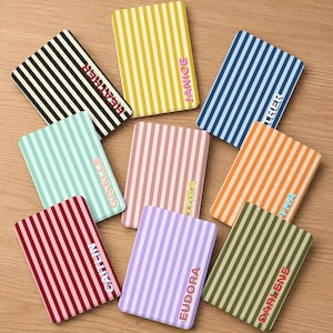 May include: A collection of personalized notebooks with vertical striped patterns in various colors, including black and white, yellow, blue, orange, green, red, pink, and purple. Each notebook features a name in a complementary color.