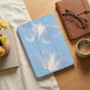 May include: A light blue tablet case with a floral design. The case is open, revealing the interior. The image also shows a brown leather journal, eyeglasses, a cup of coffee, and a small vase with yellow flowers.