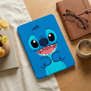 May include: A blue tablet case featuring a cartoon character with large black eyes, a dark blue nose, and a wide grin with visible teeth. The case is on a wooden surface, with a notebook, glasses, and a cup of coffee nearby.